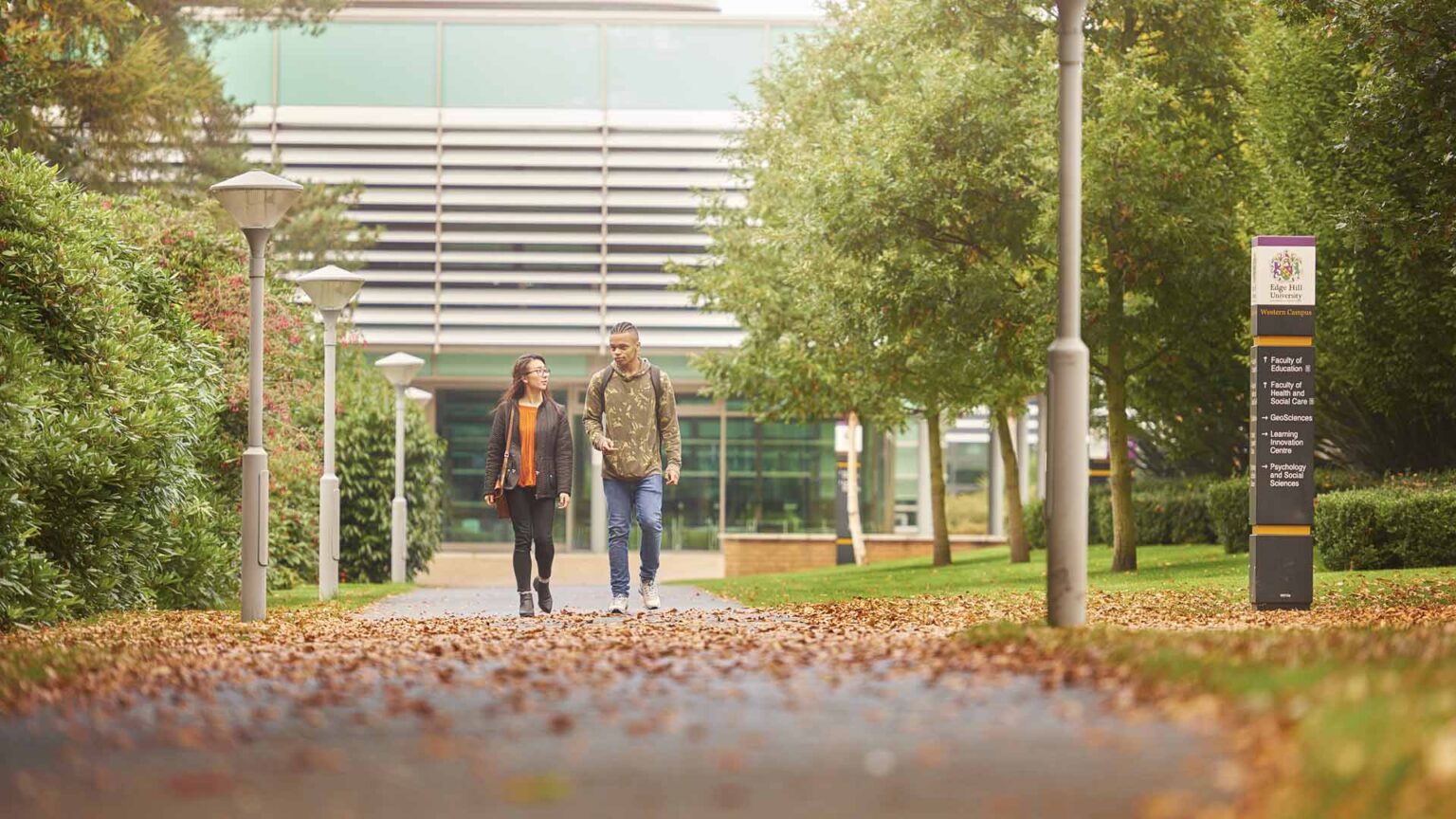 Two students walk down the pathway in front of the green Faculty of Health building on campus. Autumn leaves are on the ground and green, leafy trees line the pathway.